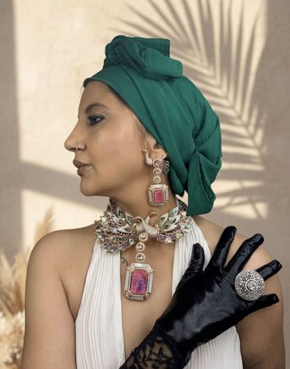Woman wearing a green headscarf, white dress, black gloves, and jewelry with pink stones.